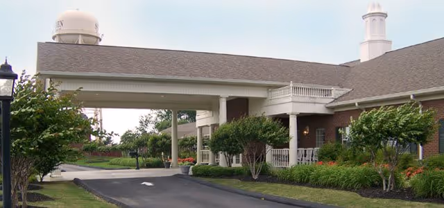 Exterior view of The Pinnacle of Southaven facility showing a covered driveway entrance with white columns, landscaped bushes and trees, and a water tower in the background under a clear sky.