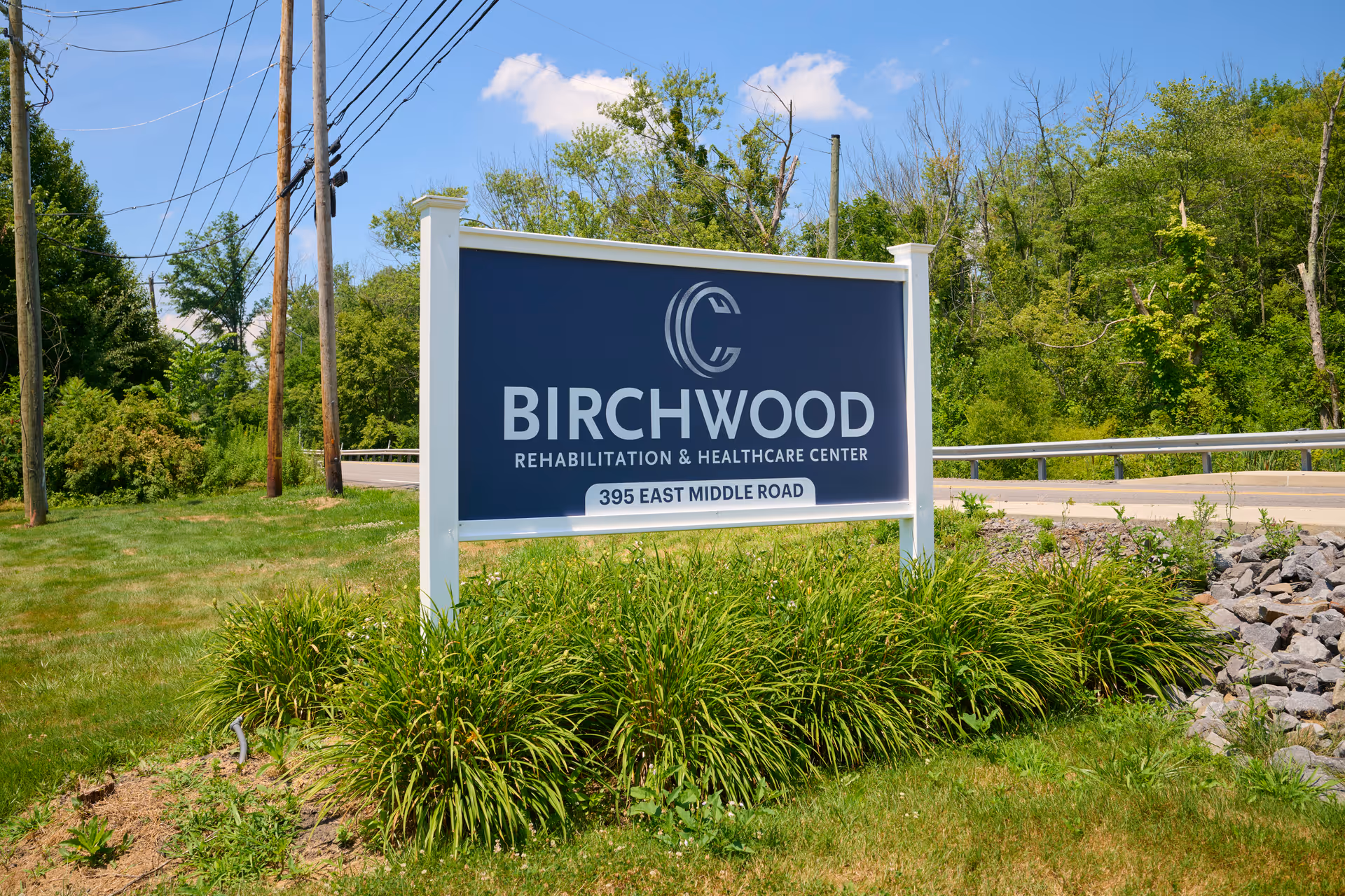 Outdoor view of a large blue and white sign for Birchwood Rehabilitation & Healthcare Center located at 395 East Middle Road, surrounded by green grass, plants, and trees under a clear blue sky.