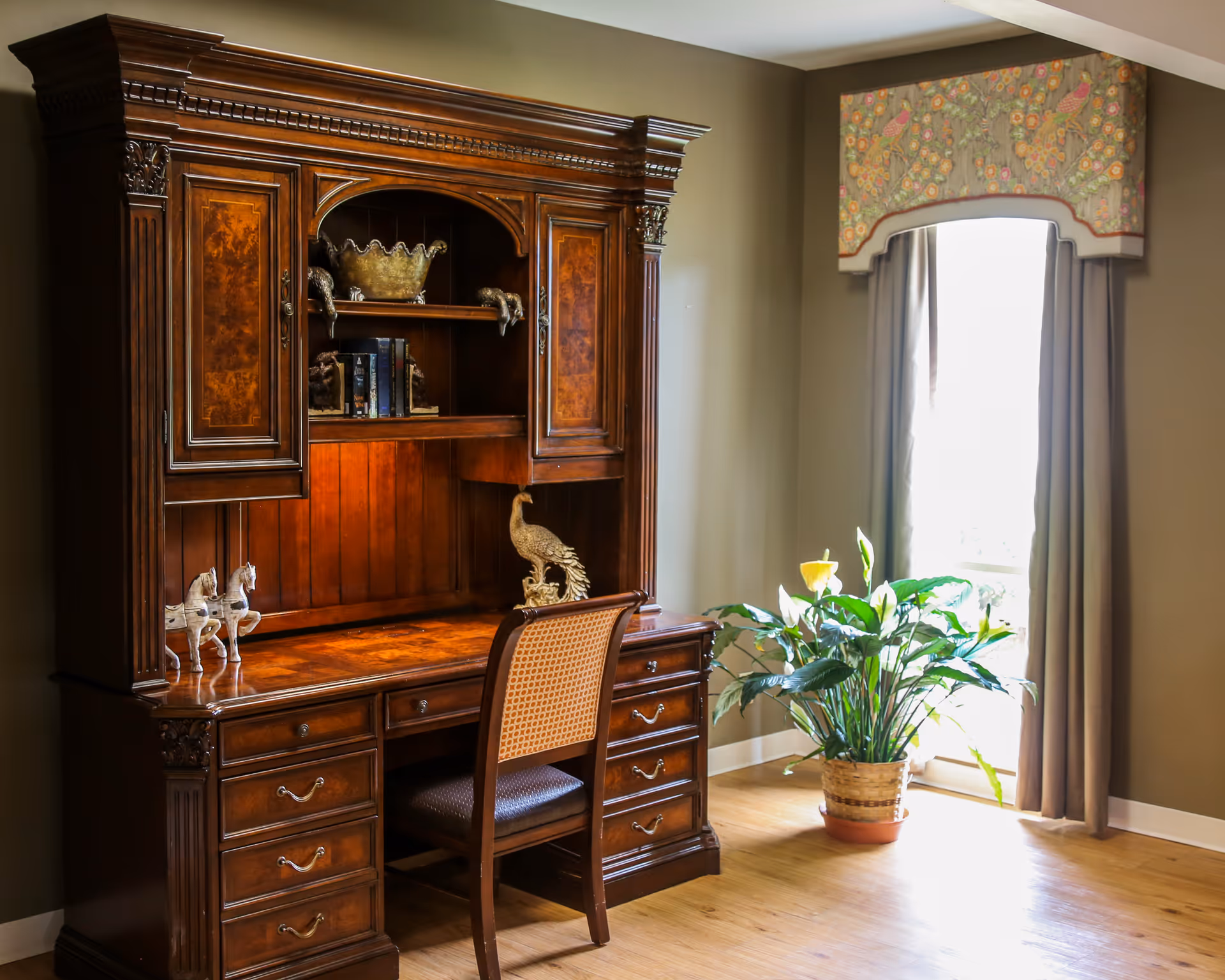 A traditional wooden desk with multiple drawers and an upper hutch featuring decorative carvings and shelves. On the desk are small horse figurines and a peacock statue. Next to the desk is a wooden chair with a woven backrest. A large potted plant sits near a window with beige curtains and a floral valance, allowing natural light to brighten the room.