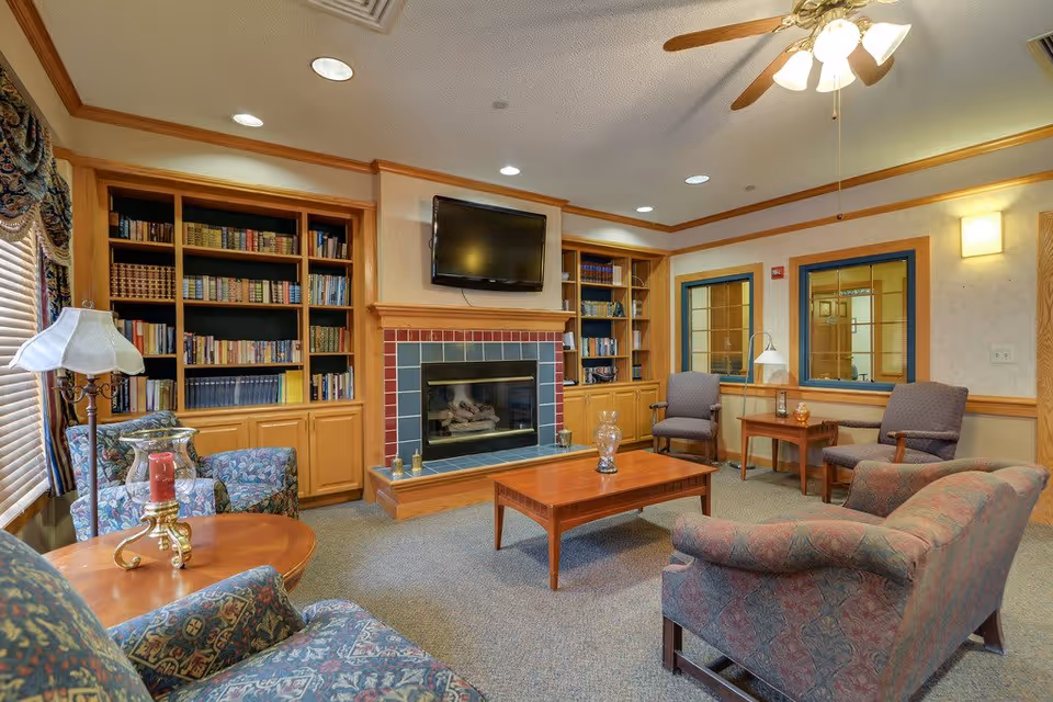 A cozy living room with patterned armchairs and a sofa arranged around a wooden coffee table. There is a fireplace with a tiled surround and a flat-screen TV mounted above it. Built-in wooden bookshelves filled with books flank the fireplace. The room has carpeted flooring, a ceiling fan with lights, and windows with wooden blinds and decorative curtains.