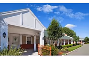Front exterior of a single-story retirement community building with a covered entrance, white columns, landscaped shrubs, and a clear blue sky.