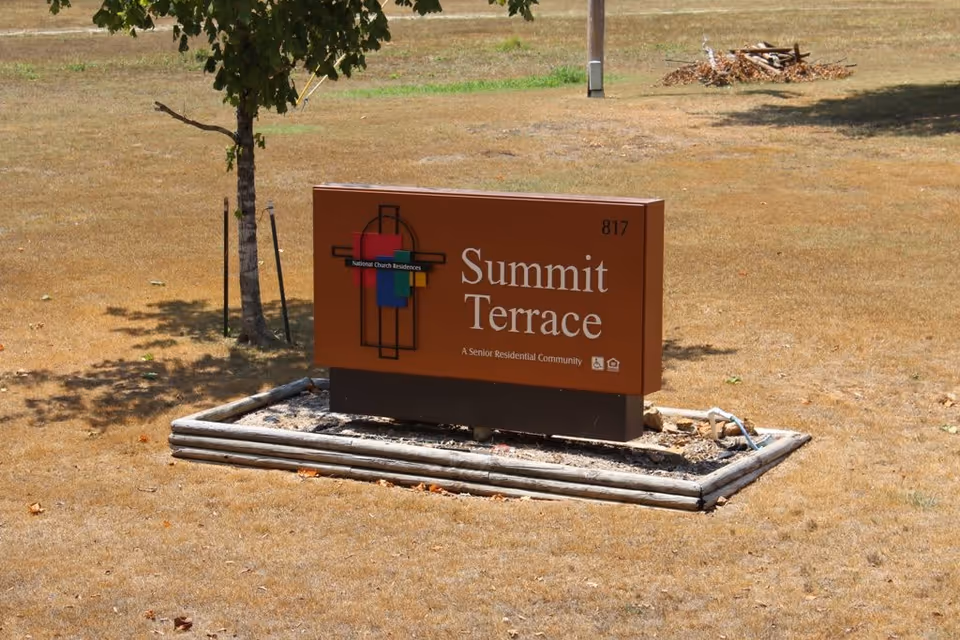 A brown sign on a patch of dry grass with a small tree nearby. The sign reads 'Summit Terrace, A Senior Residential Community' with a colorful logo and the number 817.