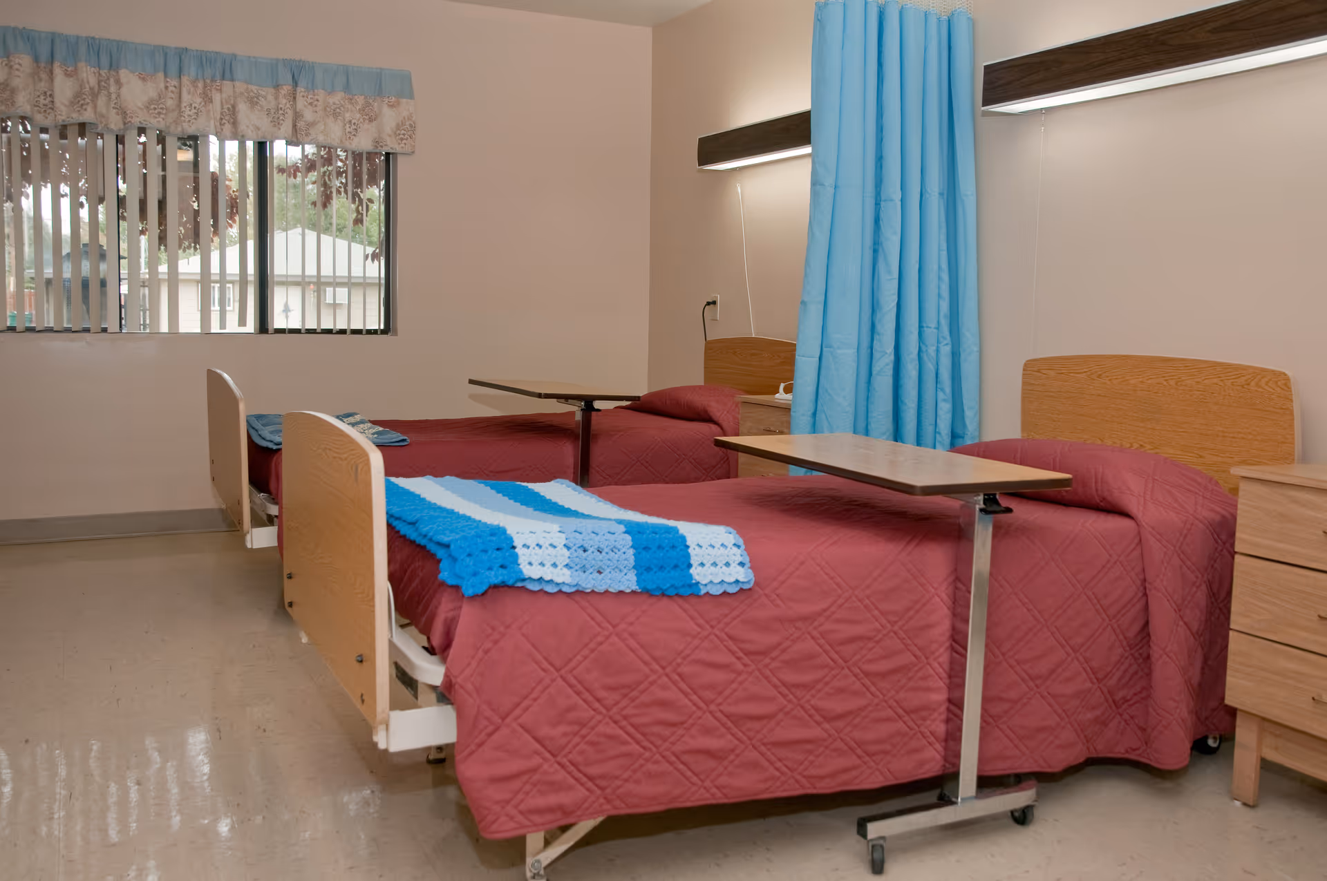 A room with two hospital-style beds covered with maroon quilts and blue and white blankets. Each bed has an overbed table and a wooden headboard. There is a blue privacy curtain between the beds, a window with vertical blinds and a valance, and a wooden dresser on the right side.