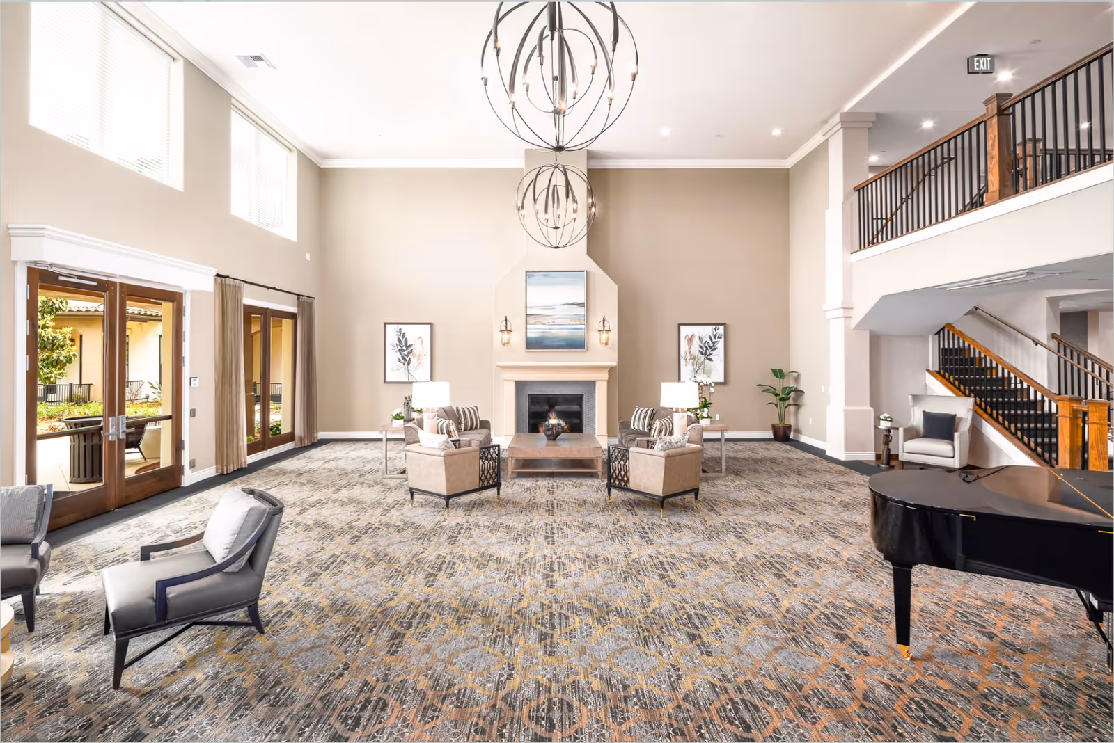 A spacious and well-lit senior living facility common area with high ceilings, large windows, and glass doors leading outside. The room features a patterned carpet, a fireplace with artwork above it, two armchairs and a coffee table in front of the fireplace, additional seating including armchairs, a grand piano, and a staircase with wooden railings leading to an upper level.
