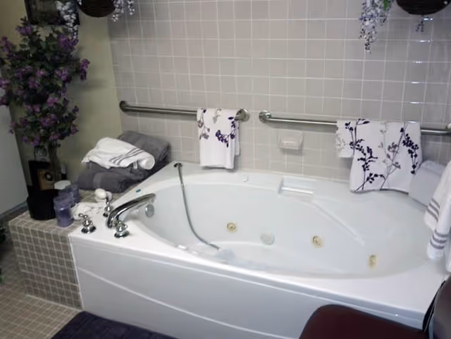 A white corner bathtub with jets in a tiled bathroom. The tub has a silver faucet and a handheld showerhead. There are two grab bars mounted on the tiled wall above the tub, each holding a white towel with purple floral designs. To the left of the tub, there is a small tiled ledge with folded towels and purple decorative items, along with a vase of purple flowers. The floor is tiled and a dark purple bath mat is partially visible.