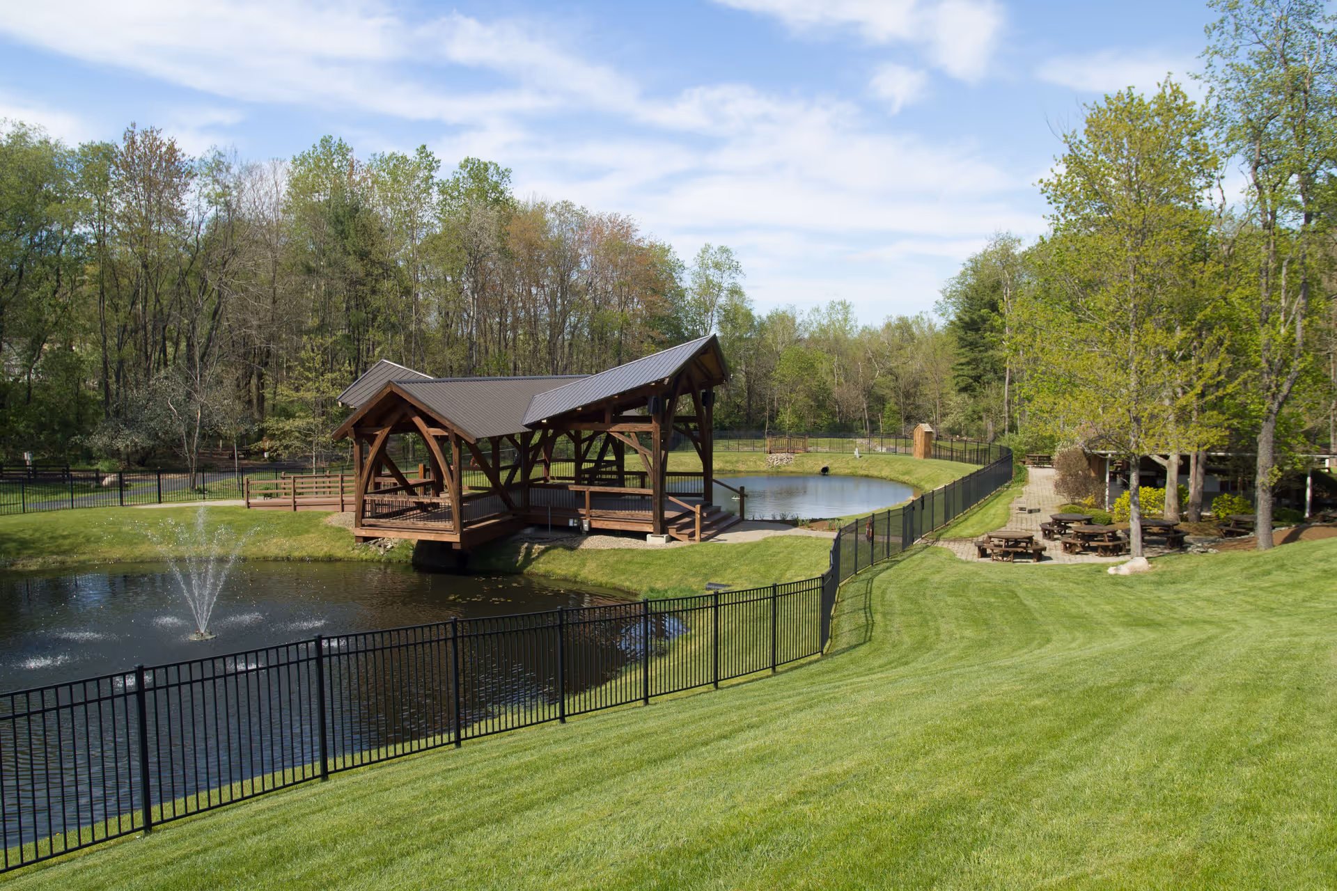 A peaceful outdoor area at Memory Garden featuring a small pond with a water fountain, a covered wooden gazebo bridge over the pond, surrounded by green grass and trees. There are picnic tables and benches along a pathway on the right side, with a forested area in the background under a partly cloudy sky.