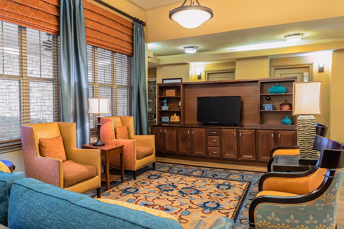 A cozy living room with a large wooden entertainment center holding a flat-screen TV, two orange armchairs with patterned cushions, a blue sofa, two wooden side tables with lamps, large windows with orange blinds and blue curtains, and a decorative area rug with blue and orange patterns.