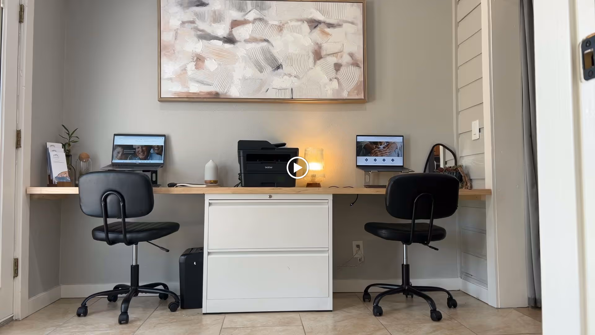 A small office workspace with a wooden desk mounted on the wall, two black swivel chairs, two computer monitors, a printer, a lamp, and some decorative items including a small plant and a framed abstract painting on the wall.