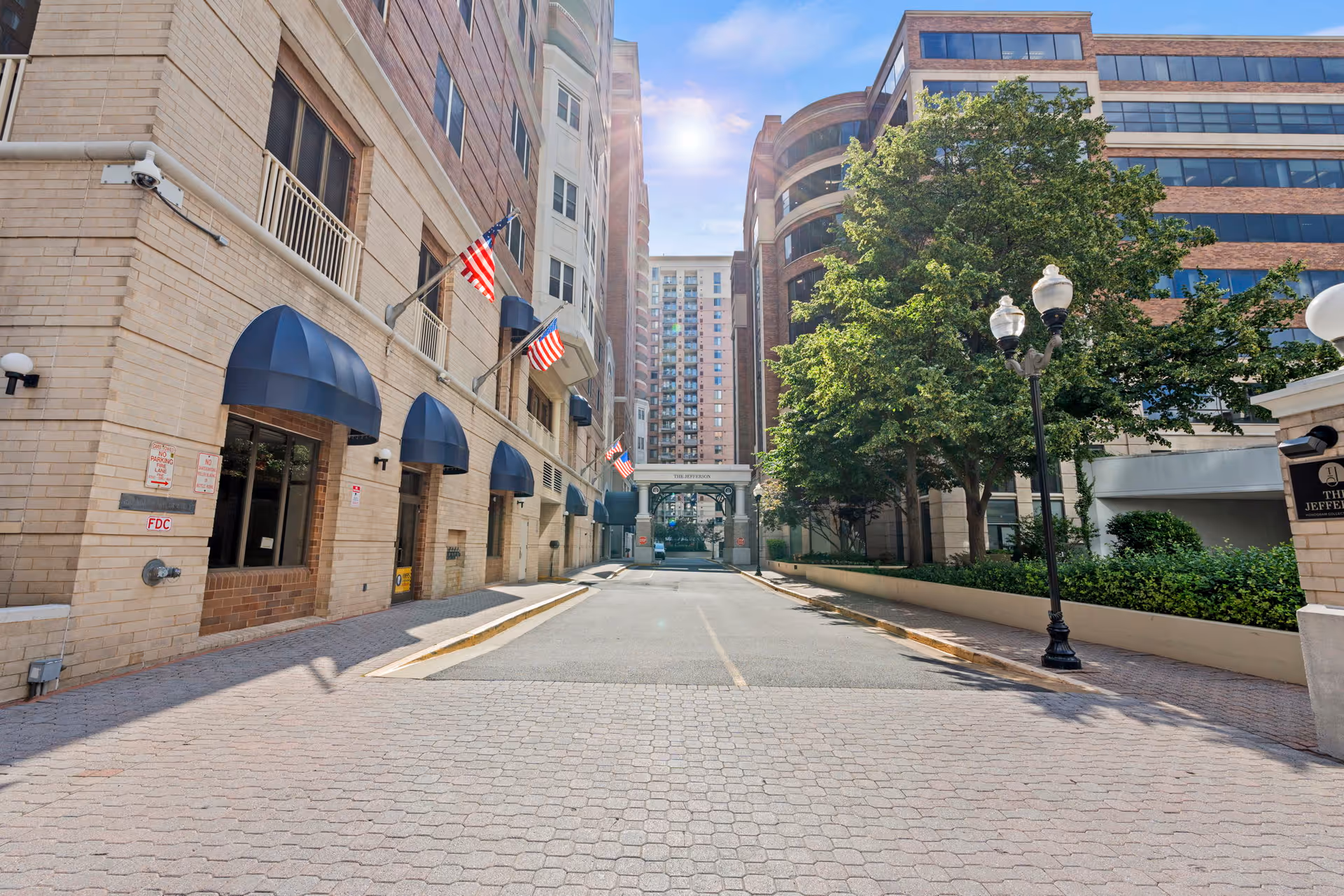 View of the exterior entrance area of The Jefferson facility, showing a paved driveway flanked by multi-story buildings with beige brick facades, blue awnings, American flags, street lamps, and trees under a partly cloudy sky.