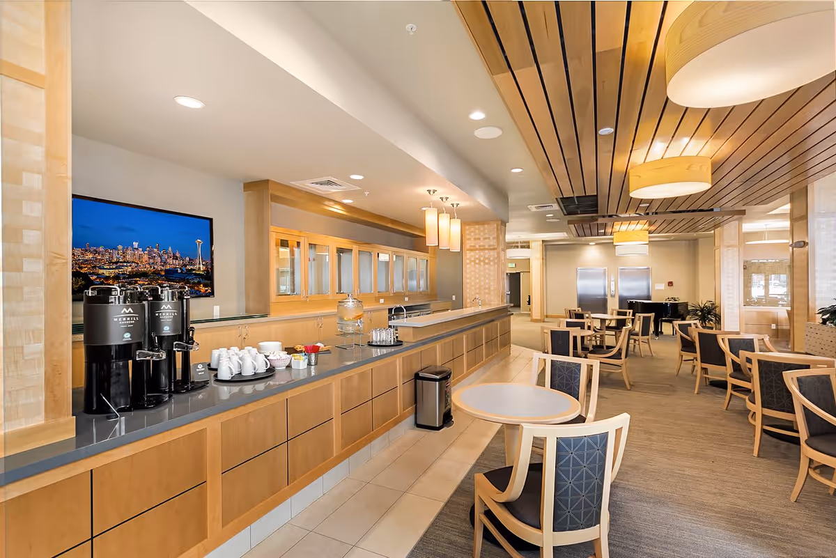 A spacious dining area with multiple round tables and wooden chairs with dark cushions. The room features a long counter with coffee dispensers, cups, and condiments. The ceiling has wooden paneling with modern hanging light fixtures. In the background, there are two stainless steel doors and a piano near the seating area.