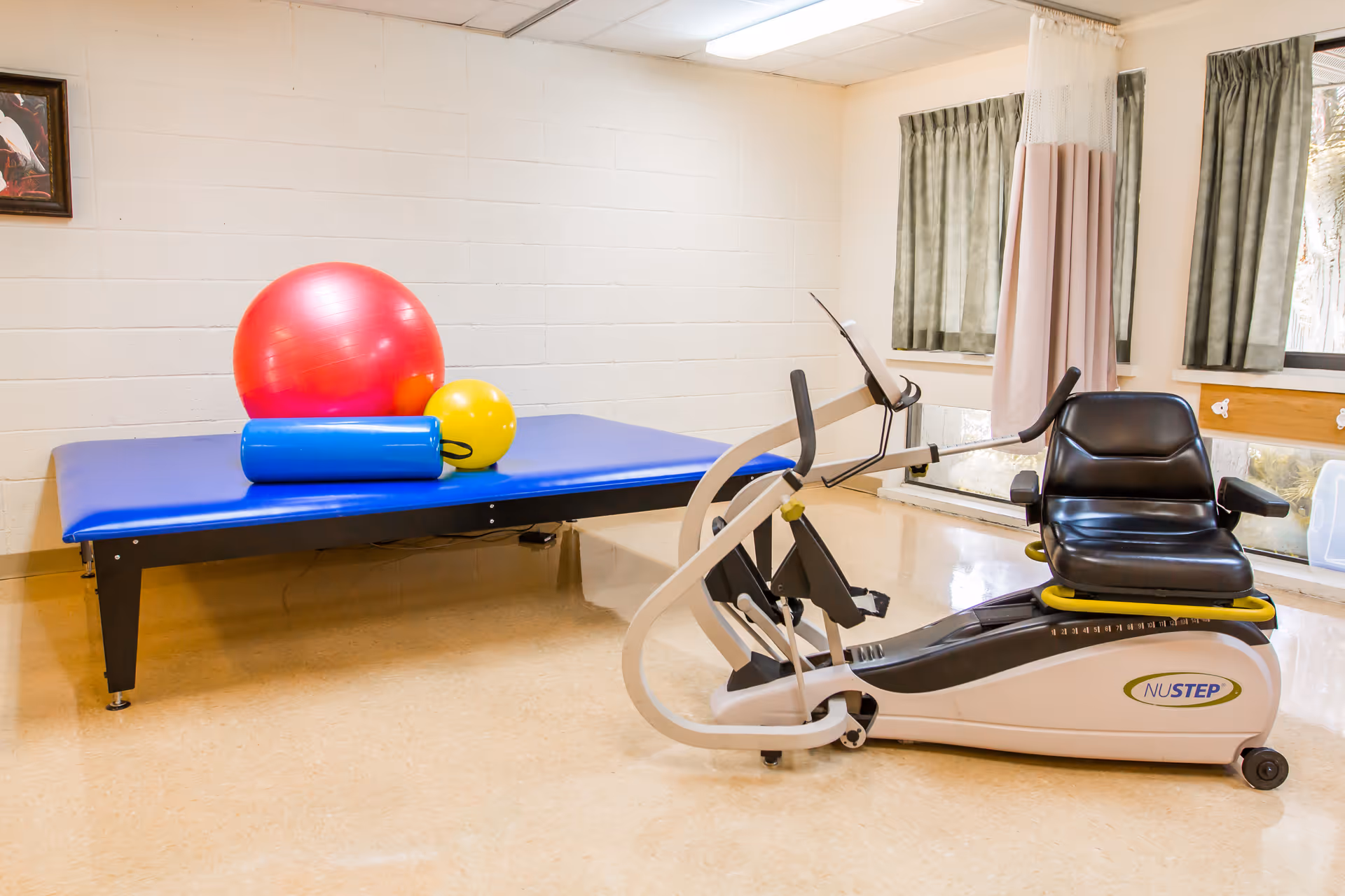 A physical therapy room with a blue padded therapy table holding a large red exercise ball, a smaller yellow ball, and a blue foam roller. Next to the table is a NuStep recumbent cross trainer exercise machine. The room has beige flooring, white walls, two windows with green curtains, and a privacy curtain.