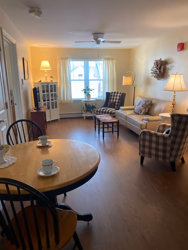 A cozy living room with a wooden round dining table and two chairs in the foreground, each with a white teacup and saucer. The room features a beige leather sofa with patterned pillows, two plaid armchairs, a small coffee table, and a TV on a wooden stand. There are two lamps providing warm light, a ceiling fan, and a window with cream-colored curtains letting in natural light. The floor is wooden, and there are some plants and decorative items around the room.