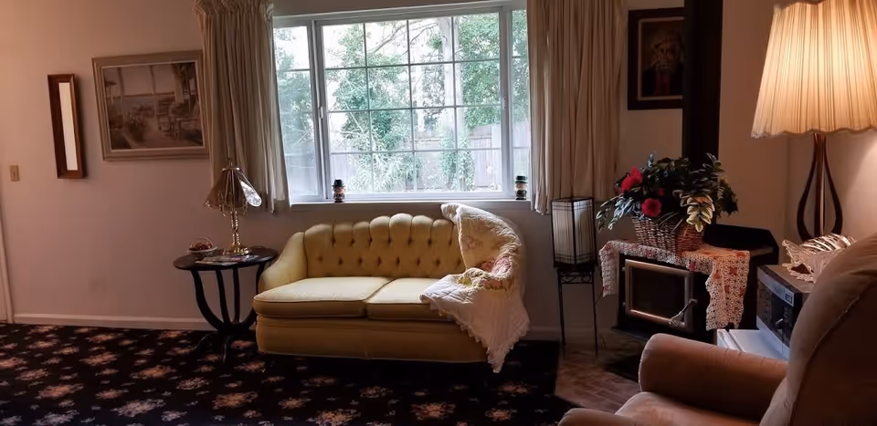 A cozy living room with a yellow tufted loveseat covered partially by a quilt, a small round side table with a lamp and decorative items, a large window with curtains showing greenery outside, a fireplace with a basket of flowers on top, a floor lamp with a pleated shade, and a comfortable armchair in the foreground.