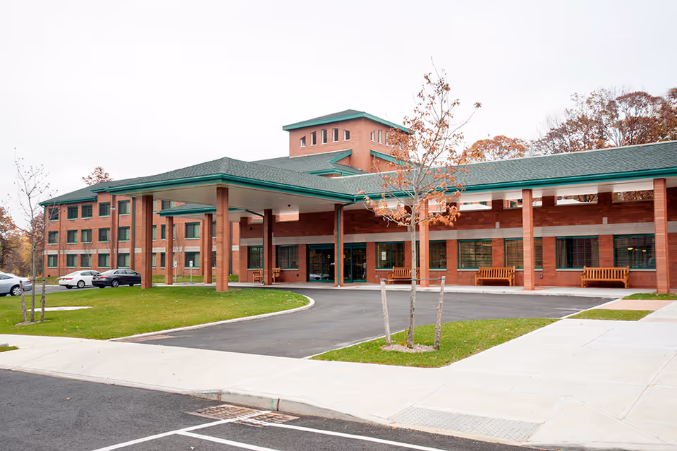 Exterior view of a large brick senior living facility with a covered entrance supported by multiple columns. There are benches along the front wall, a few small trees with autumn leaves, and several parked cars visible on the left side. The sky is overcast.