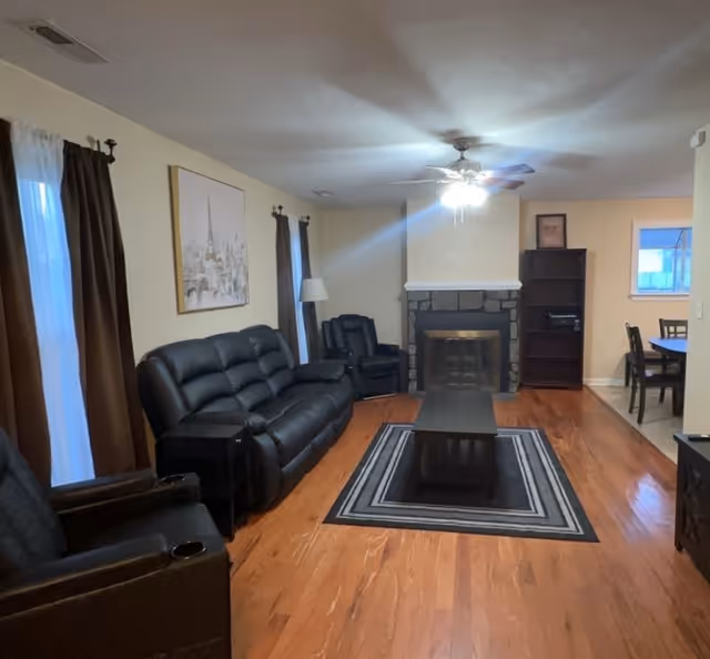 Spacious living room with leather sofas and recliners arranged around a coffee table and a stone fireplace under a ceiling fan.