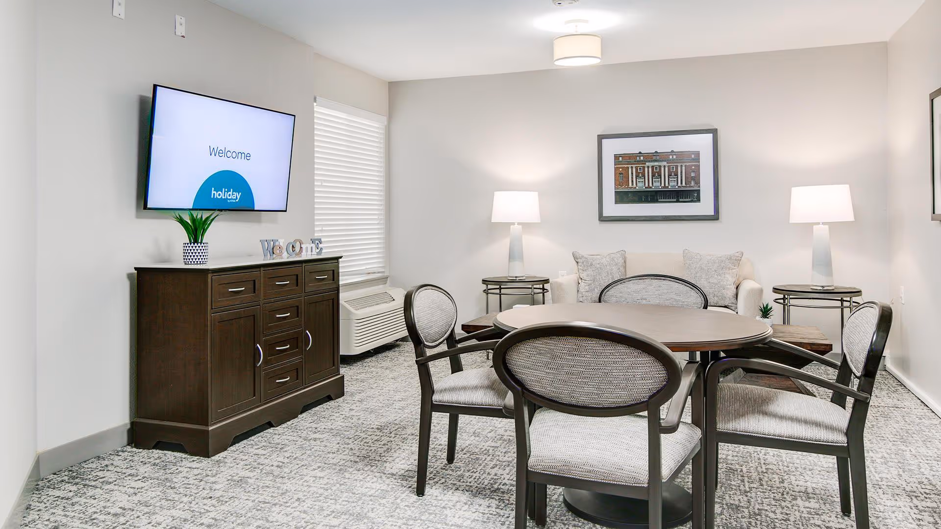 A cozy living room area with a round table surrounded by four upholstered chairs. In the background, there is a beige sofa with two cushions, flanked by two side tables each holding a lamp. A framed picture hangs on the wall above the sofa. On the left side, a dark wooden cabinet holds a small plant and decorative letters spelling 'Welcome'. A flat-screen TV is mounted on the wall above the cabinet displaying a welcome message.