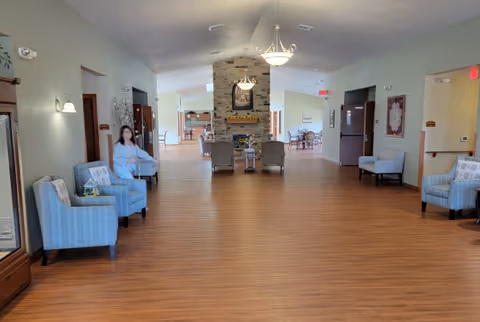 A spacious senior living facility common area with wooden flooring, light green walls, and several blue upholstered chairs arranged along the walls. In the center background, there is a stone fireplace with a framed picture above it and two armchairs facing the fireplace. Ceiling lights hang from the vaulted ceiling, and a woman is sitting on one of the chairs on the left side.