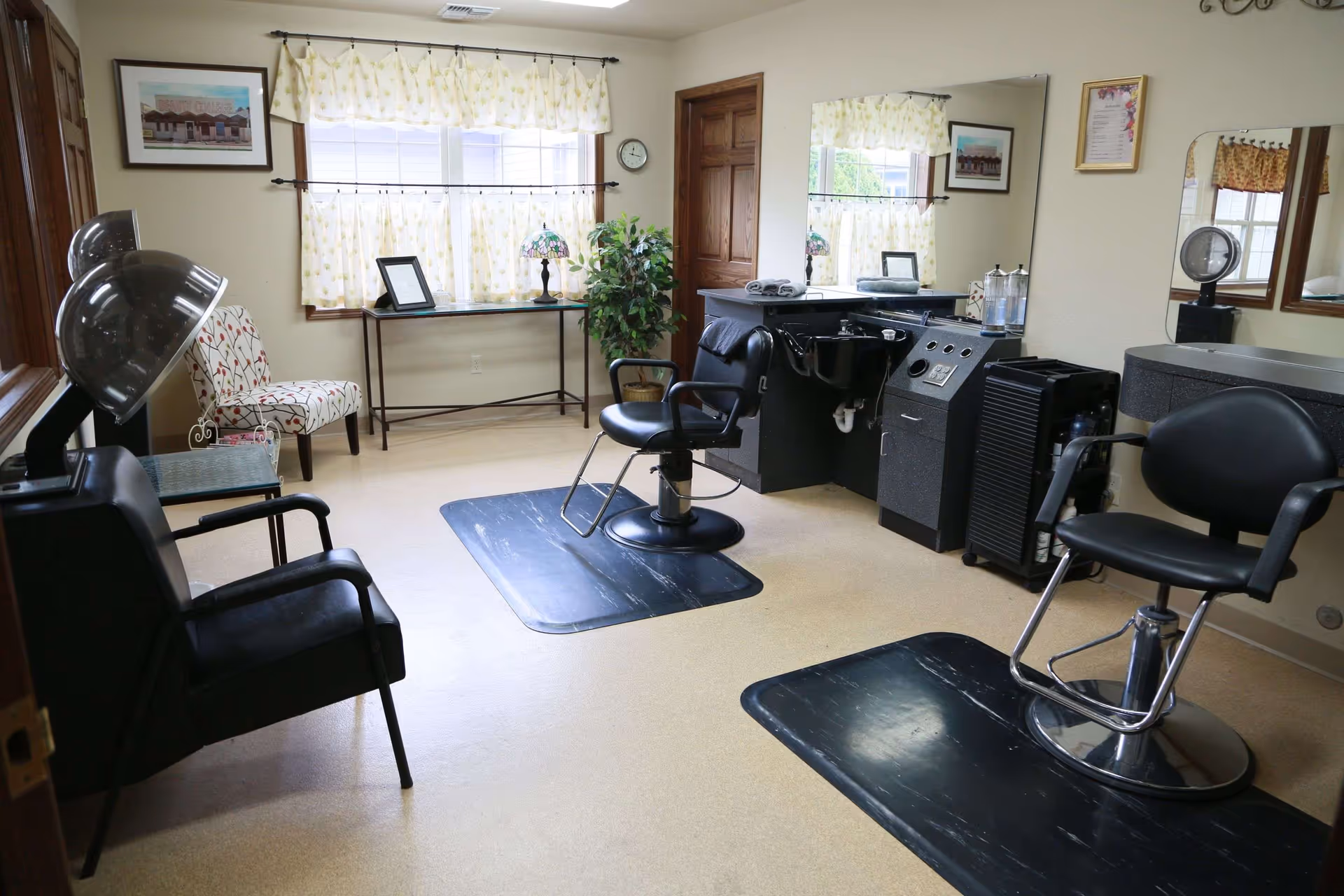 Interior of a hair salon room with two black salon chairs on black mats, a hair washing station, a hair dryer chair, a patterned armchair, a small table with a lamp, and windows with yellow patterned curtains.