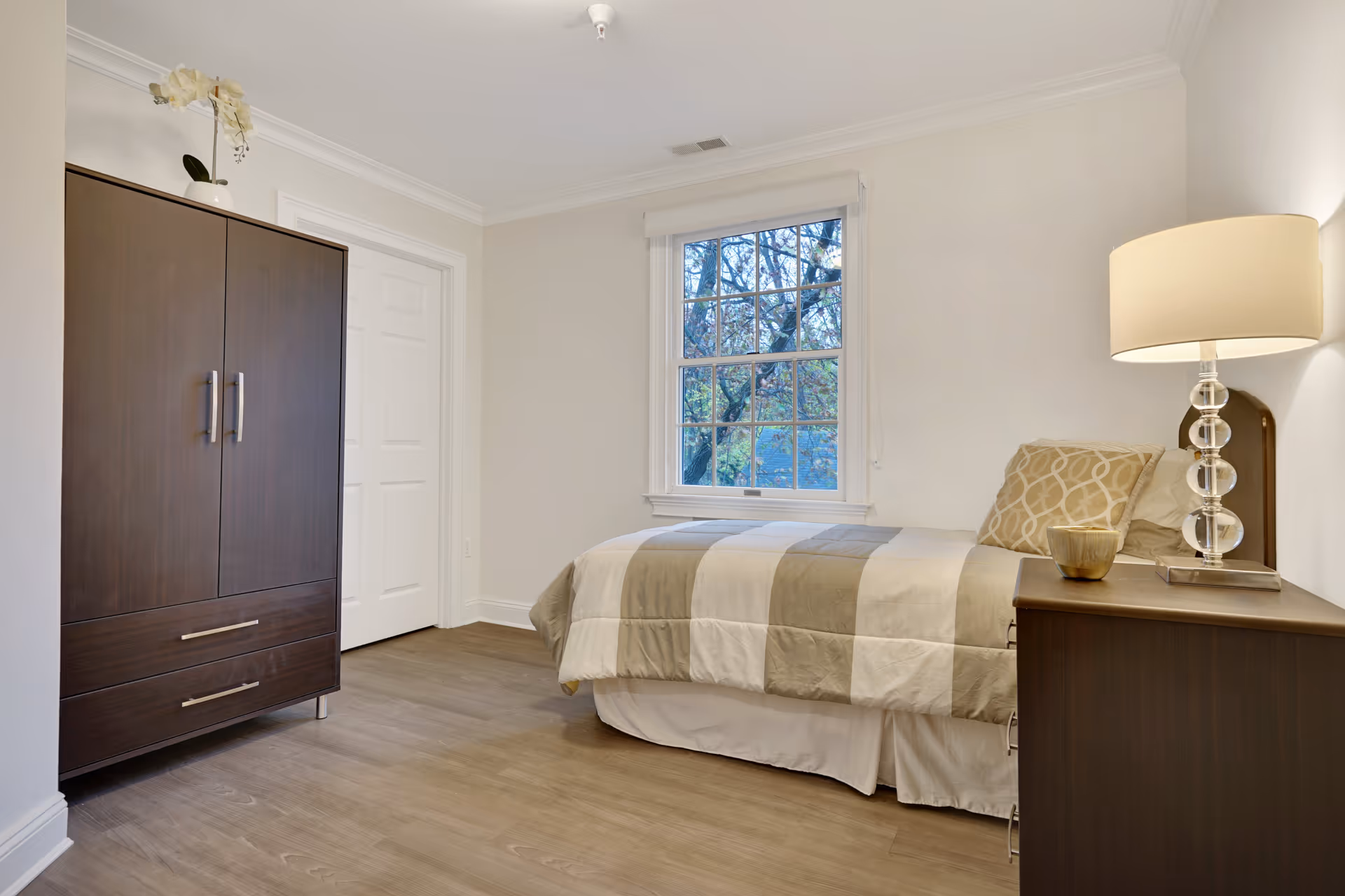 A simple, clean bedroom with a single bed covered in a beige and white striped comforter, a wooden nightstand with a lamp and a cup, a large window showing trees outside, and a dark wooden wardrobe with drawers. The room has light-colored walls and wood flooring.