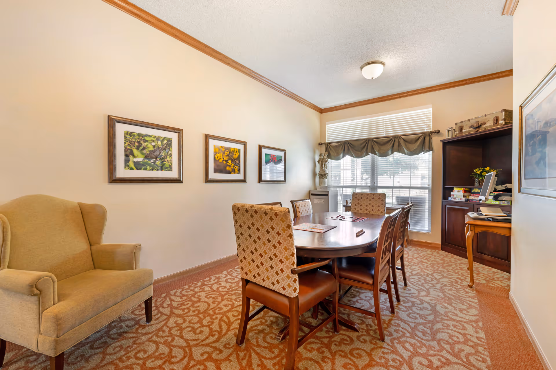 Bright dining/meeting room with an oval table and chairs, an upholstered armchair, framed artwork on the wall, and a bookshelf by a large window.