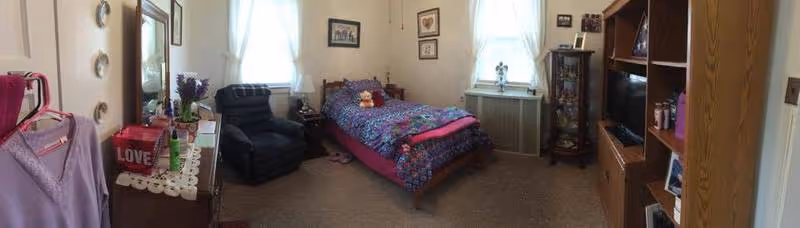 A cozy bedroom with a single bed covered in a colorful floral quilt and a pink blanket. The room has two windows with white curtains, a dark recliner chair, a wooden dresser with a mirror, and various decorative items including framed pictures on the walls and a small cabinet with glass doors displaying collectibles. There is also a wooden shelving unit with books and other items.