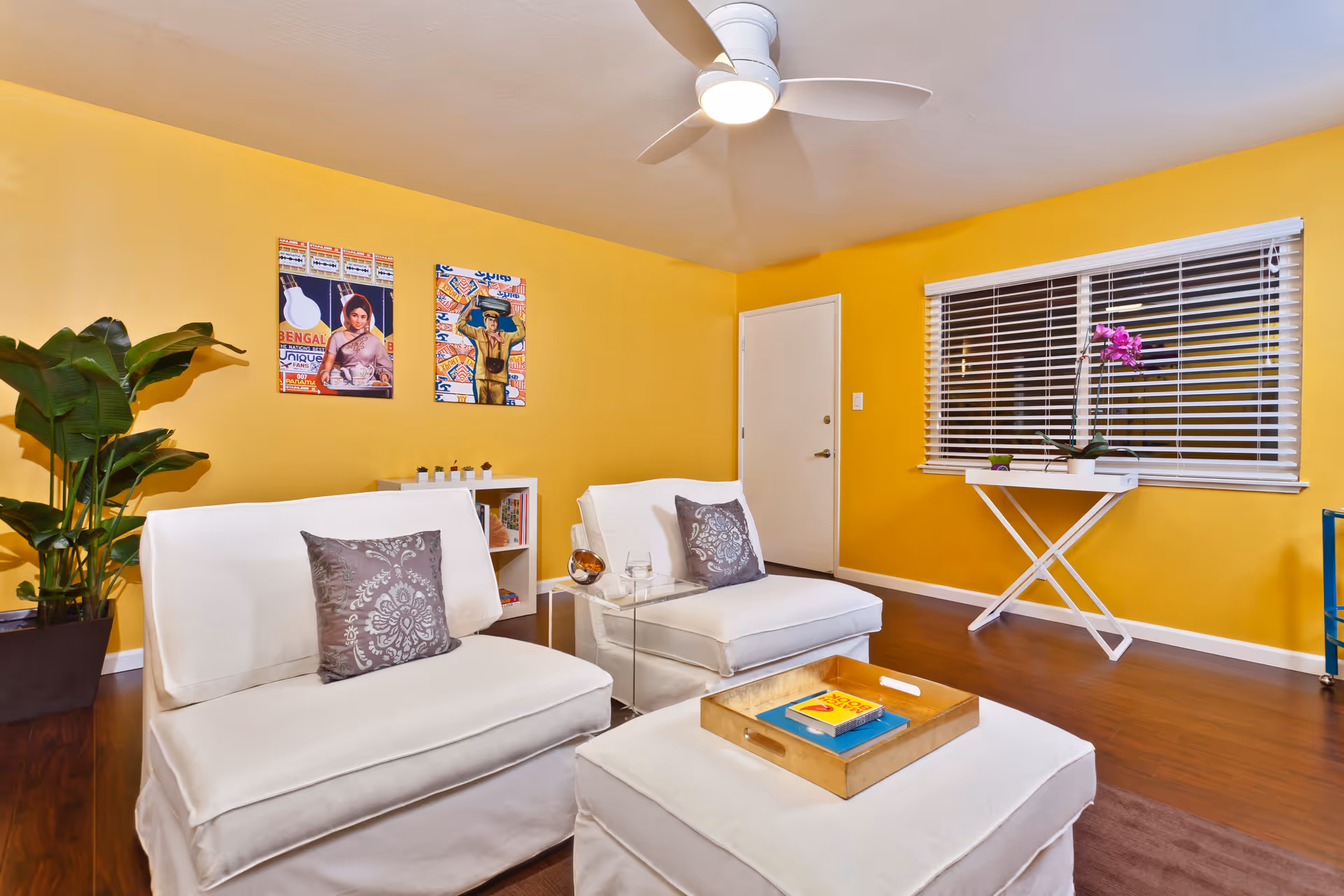 A cozy living room with bright yellow walls, two white cushioned chairs with decorative pillows, a matching ottoman with a wooden tray holding books, a small white table with a purple orchid near a window with white blinds, two colorful framed artworks on the wall, a green potted plant, and a ceiling fan with light.