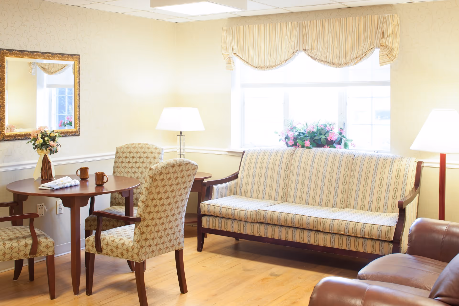 Bright common room with a striped sofa, upholstered chairs around a small round table, lamps, and a window with flowers.