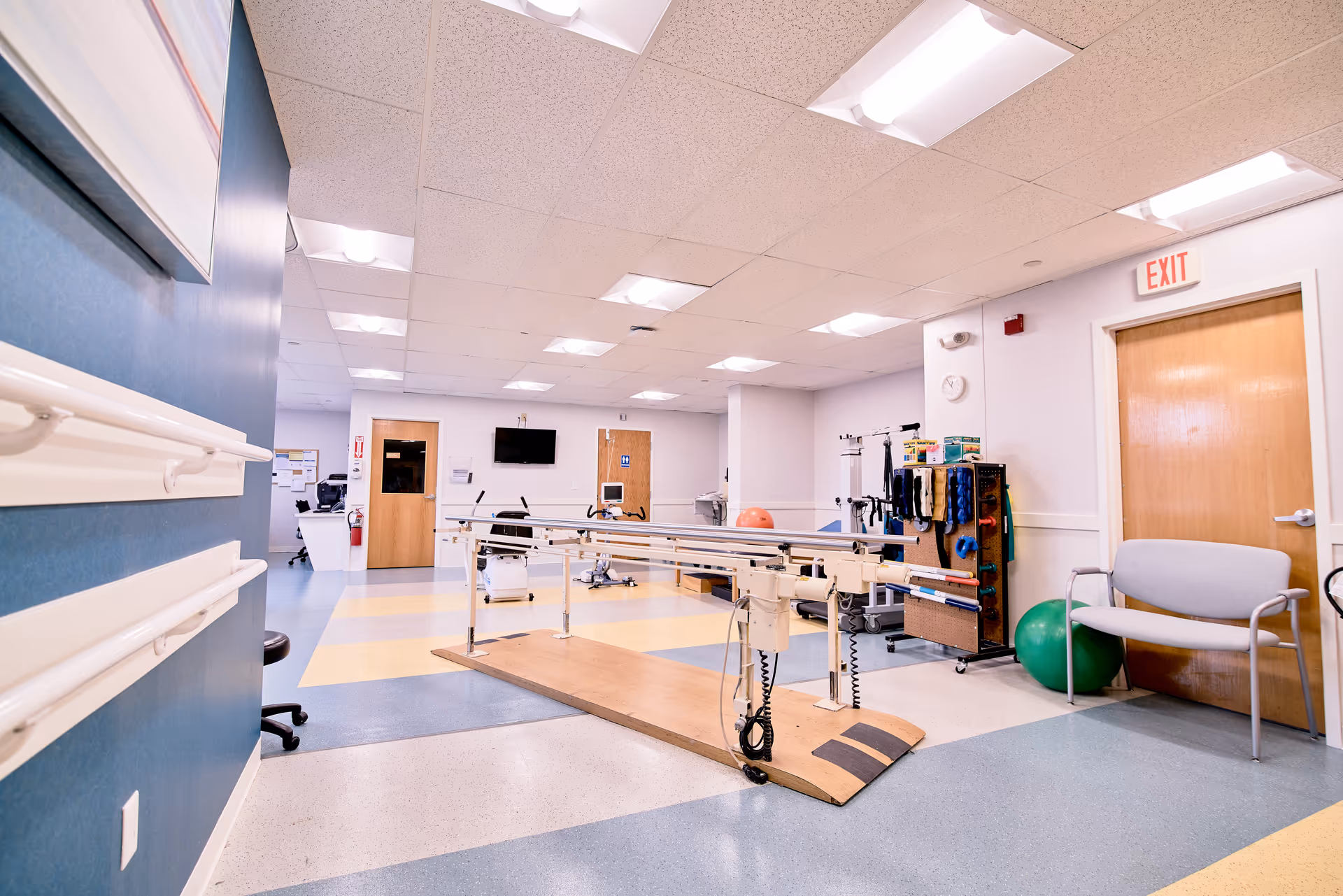 A rehabilitation or physical therapy room in a senior living facility with parallel bars for walking exercises, exercise equipment, a chair, and a green exercise ball. The room has a blue and yellow patterned floor, white walls, and fluorescent ceiling lights. There are doors, a wall-mounted TV, and handrails along one wall.