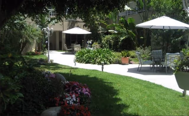 Sunlit courtyard with a green lawn, flower beds, paved walkways, and patio tables with umbrellas surrounded by building greenery.
