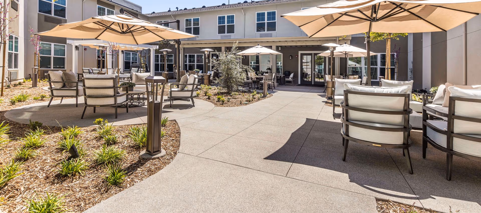 Sunny courtyard with patio tables, umbrellas, and cushioned chairs in front of a two-story senior living building.
