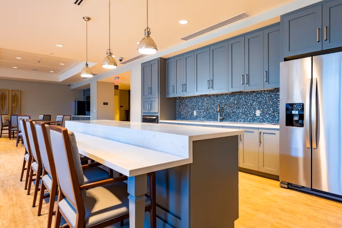 Modern kitchen area with a large white marble island countertop surrounded by wooden chairs with cushions. The kitchen features blue-gray cabinets, a mosaic tile backsplash, stainless steel refrigerator, and three hanging pendant lights above the island. In the background, there is a dining area with tables and chairs.