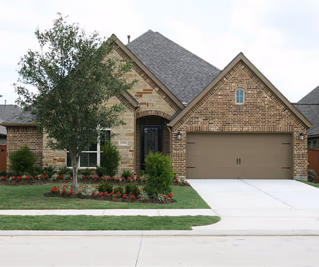 Front exterior view of a single-story brick house with a two-car garage, a tree, and landscaped flower beds in the front yard.