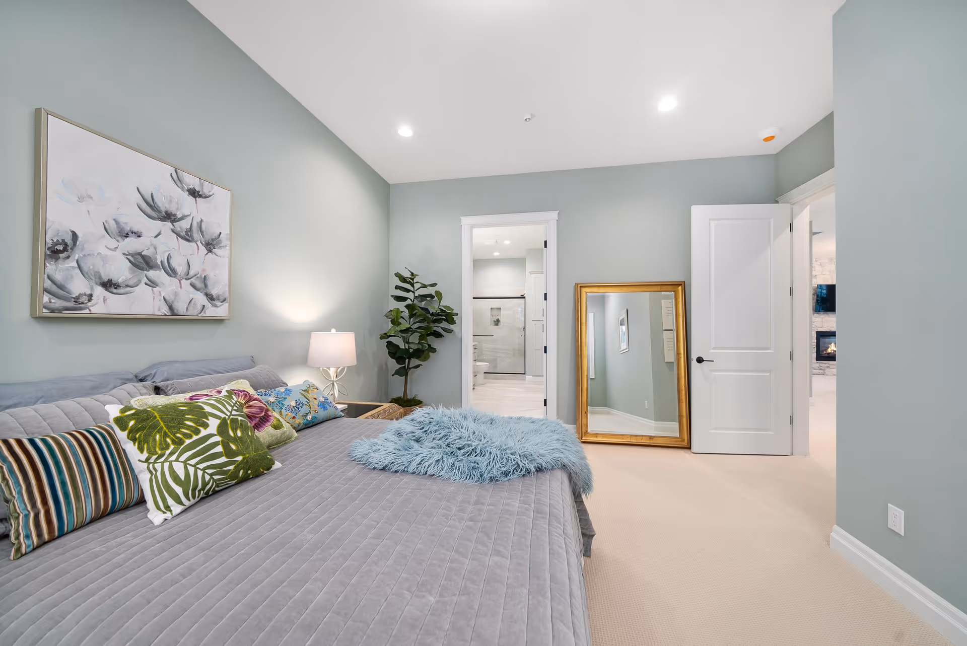 A bedroom with a large bed covered in a gray quilt and colorful pillows including striped and leaf-patterned designs. A bedside table with a lamp and a potted plant are next to the bed. A large framed mirror leans against the wall near an open door leading to a bathroom with a glass shower. The walls are painted light gray and the floor is carpeted in beige.