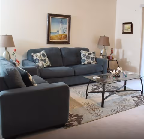 A cozy living room with two dark gray sofas adorned with patterned cushions, a glass coffee table with decorative items, two table lamps on side tables, a floral area rug, and framed artwork on the beige walls.