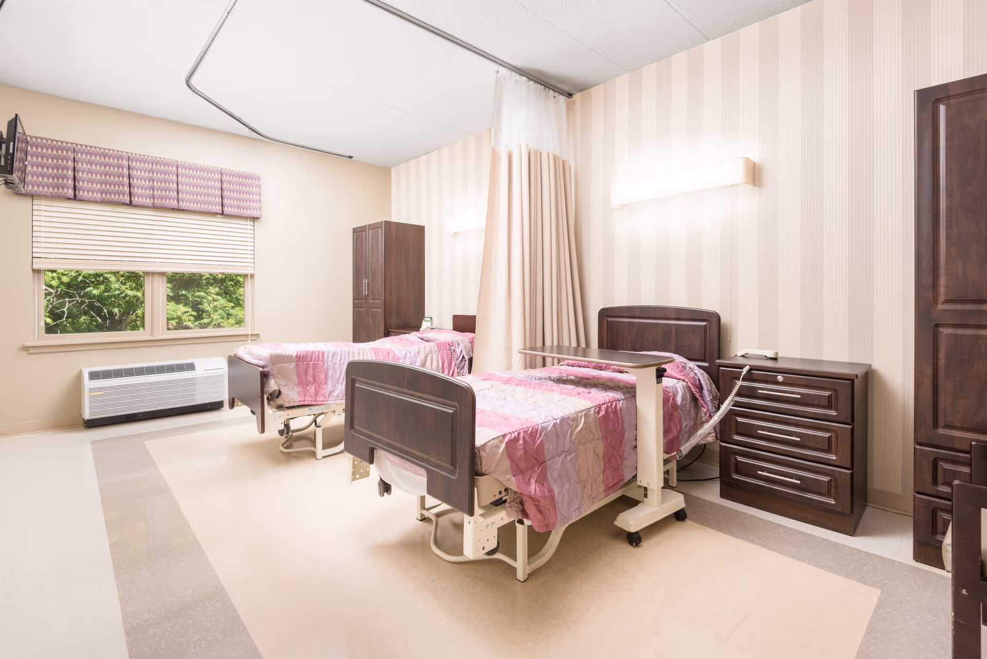 A bright and clean room with two hospital-style beds covered in pink and purple patterned bedspreads. Each bed has a dark wooden headboard and footboard, and there is a dark wooden nightstand with drawers between the beds. A privacy curtain hangs from the ceiling between the beds. The room has beige walls with vertical stripes and a window with blinds and a purple valance, showing green trees outside. There is an air conditioning unit below the window and a tall dark wooden cabinet in the corner.
