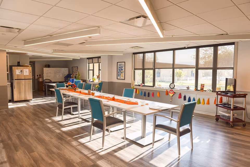 A bright, spacious room with large windows letting in natural light. The room features a long white table with teal chairs arranged around it. The table is decorated with small pumpkins and a festive orange runner. The floor is wood-style laminate, and there is a stainless steel refrigerator in the background. Colorful tassel garlands hang below the windows, and a small cart with a TV is positioned near the window.