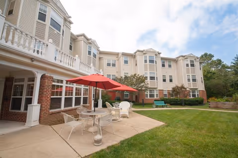 Outdoor patio area of a senior living facility with white metal chairs and tables, one table shaded by a red umbrella, adjacent to a multi-story beige building with white trim and large windows. The area is surrounded by green grass and some trees in the background under a partly cloudy sky.