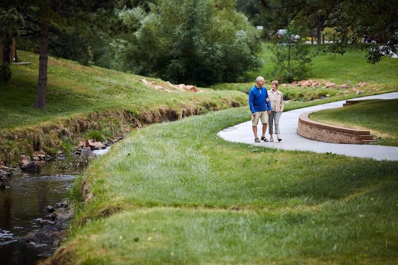 An elderly couple walking arm in arm on a paved path beside a small creek in a lush green park-like setting with trees and grass.