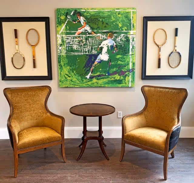 Two vintage-style armchairs with wooden legs and yellow upholstery are positioned on either side of a small round wooden table. Above the table hangs a colorful painting depicting two tennis players in action on a green court. On each side of the painting, there are framed displays of three vintage wooden tennis rackets mounted on a beige background. The setting appears to be an interior room with wooden flooring and a light-colored wall.