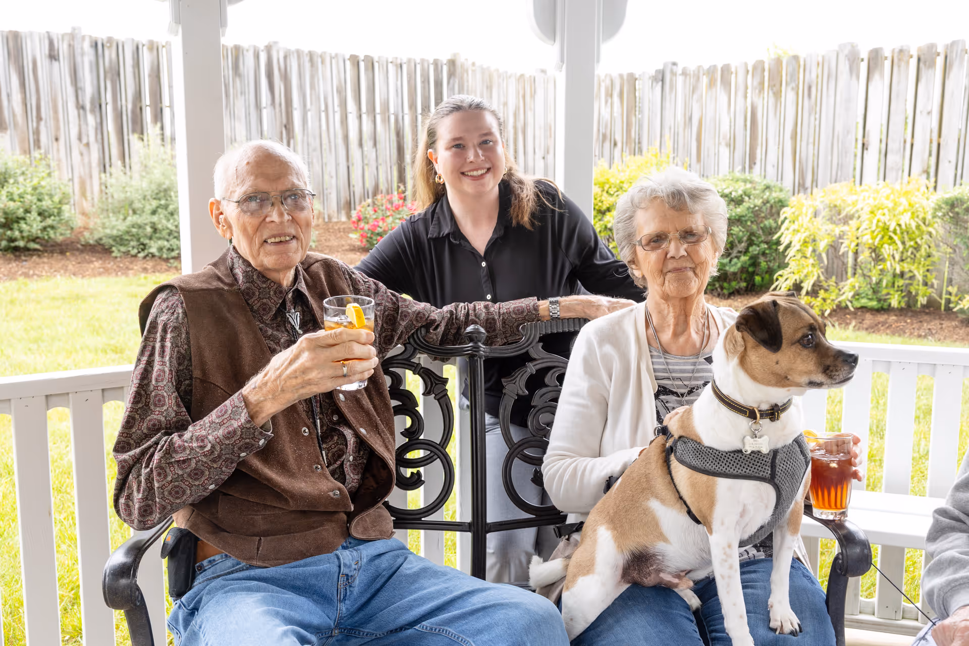 An elderly man and woman sitting on a bench outside on a porch with a wooden fence and greenery in the background. The man is holding a glass of iced tea with a lemon slice, and the woman is holding a small dog wearing a harness. A younger woman stands behind them, smiling.
