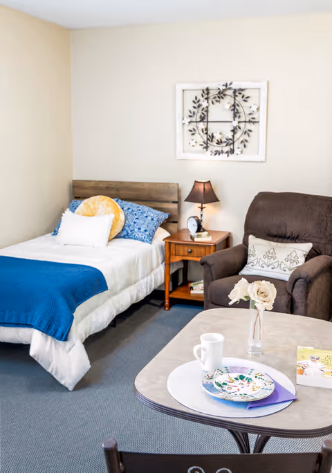 A cozy bedroom in a senior living facility featuring a single bed with white bedding, blue blanket, and decorative pillows. Next to the bed is a wooden nightstand with a lamp, clock, and books. A comfortable brown armchair with a patterned pillow is placed beside the nightstand. In the foreground, there is a small table set with a floral plate, white mug, purple napkin, and a vase with white flowers. The walls are light-colored with a decorative wall hanging above the nightstand.