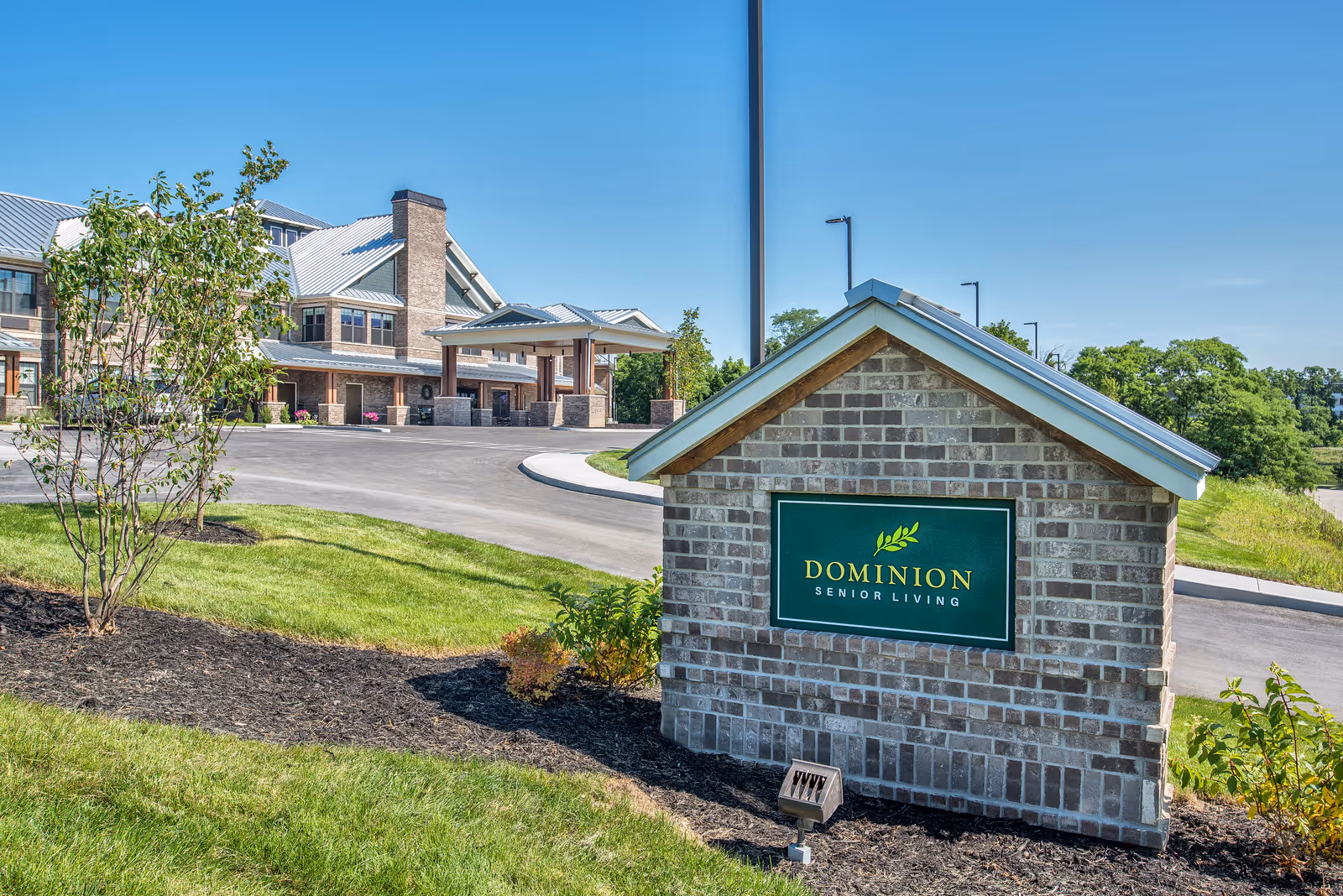 Exterior view of Dominion Senior Living of Florence facility with a brick sign in the foreground and the building with a covered entrance in the background under a clear blue sky.