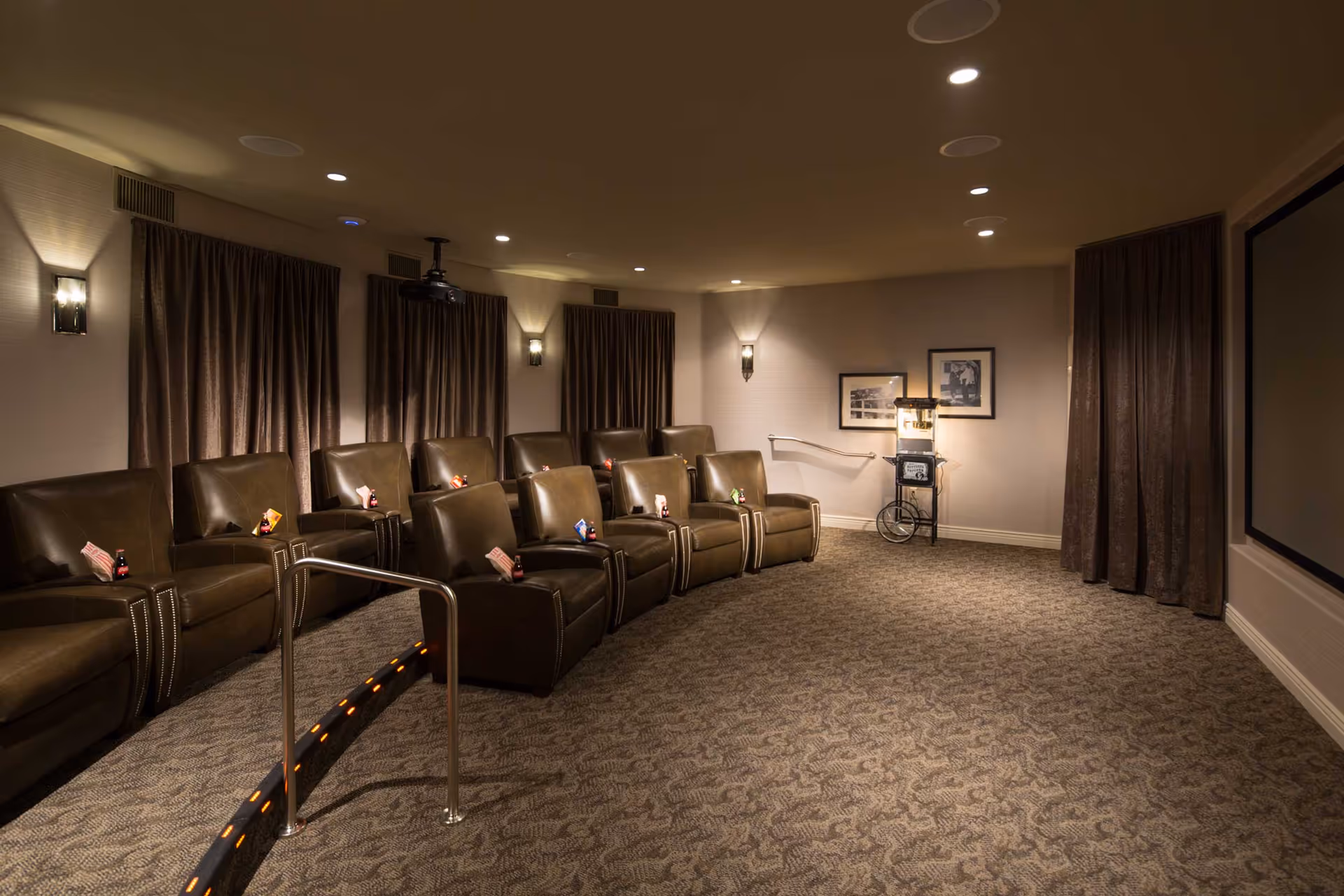 A cozy private theater room with two rows of brown leather recliner chairs, each with a small bottle of soda and a bag of popcorn. The room has dim lighting with wall sconces, dark curtains covering the windows, a popcorn machine in the corner, and a large projection screen on the right wall.