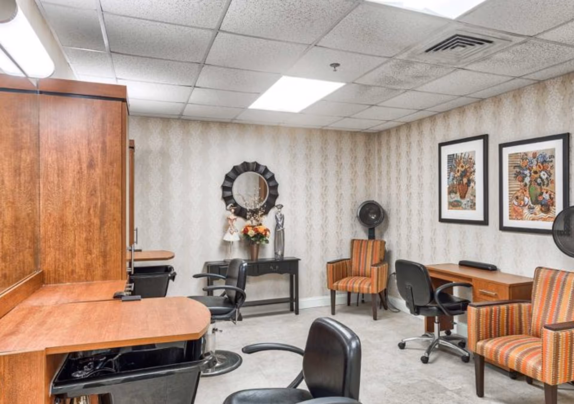 Interior of a senior living facility room set up as a salon with wooden counters, black salon chairs, two striped armchairs, a small black table with decorative items and a round mirror, two framed floral paintings on the wall, and a hair dryer hood in the corner.