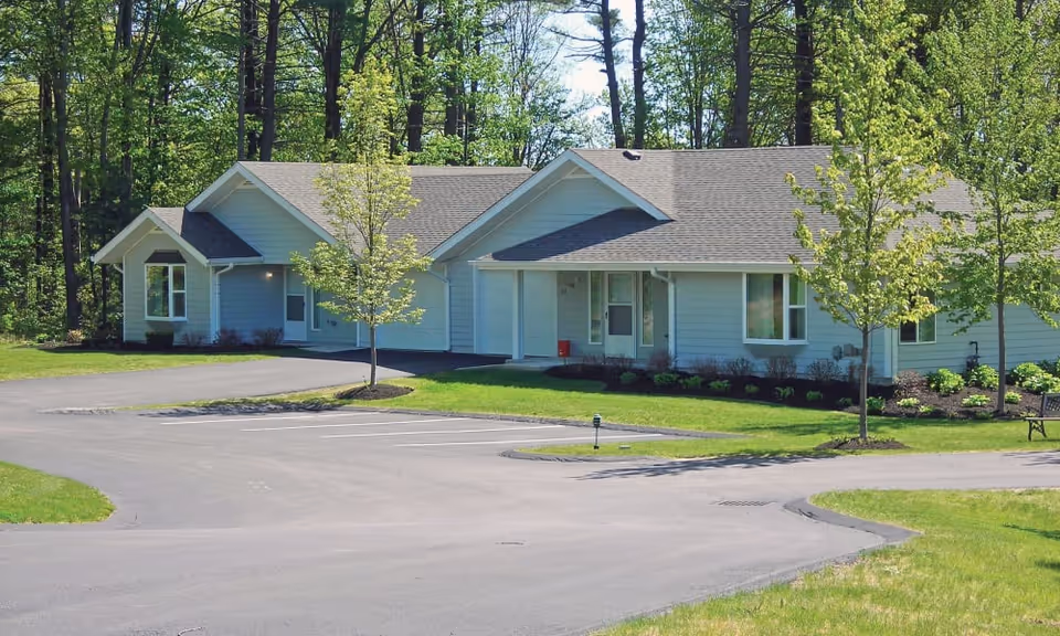 Single-story light-blue senior living building with a paved driveway, lawn, and young trees in front.