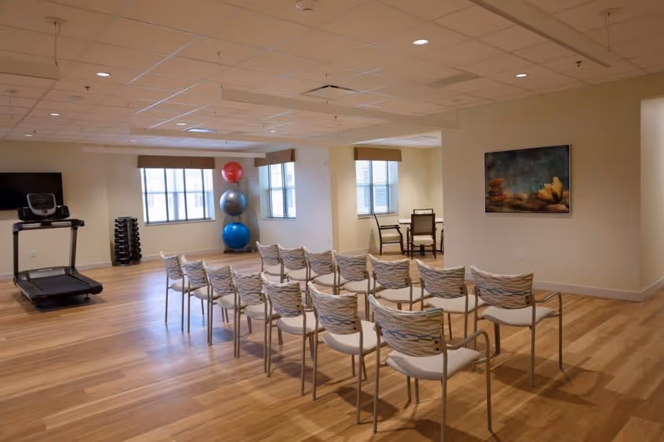 A spacious room with wooden flooring featuring two rows of chairs arranged facing a blank wall. To the left, there is a treadmill, a rack of dumbbells, and three large exercise balls stacked vertically near two windows. In the background, there is a small table with four chairs near another window. A framed picture hangs on the right wall.