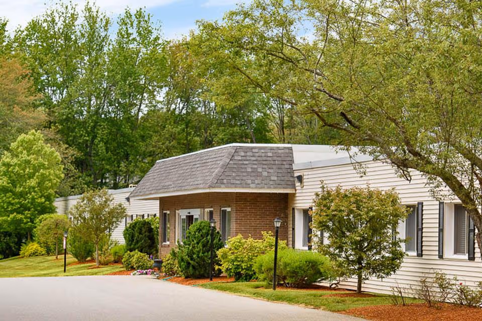 Exterior view of a single-story senior living facility building surrounded by green trees and shrubs, with a paved driveway in front and a partly cloudy sky above.