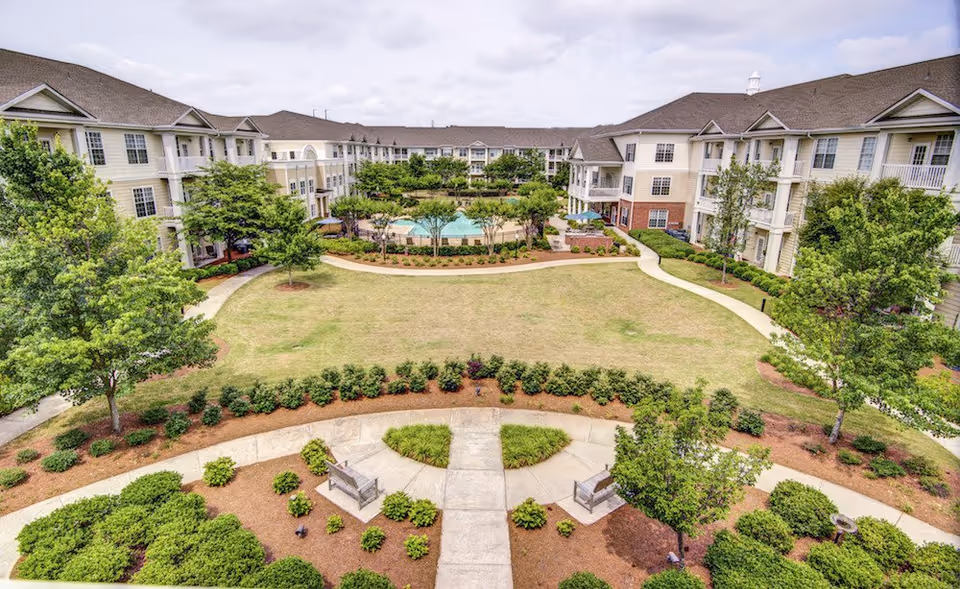 View of a senior living facility courtyard with well-maintained landscaping, walking paths, benches, and a swimming pool surrounded by three-story residential buildings under a cloudy sky.