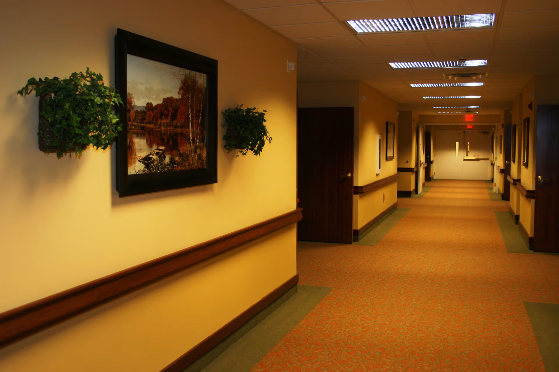 A long, well-lit hallway in a nursing center with yellow walls, brown handrails, and patterned carpet. The walls are decorated with framed landscape paintings and green potted plants. Several closed wooden doors line the hallway, and double exit doors are visible at the far end.