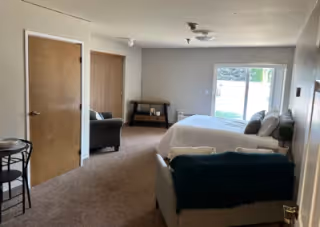 Spacious senior-living studio room with a bed, sofa, armchair, small table, and sliding glass door to the outside.